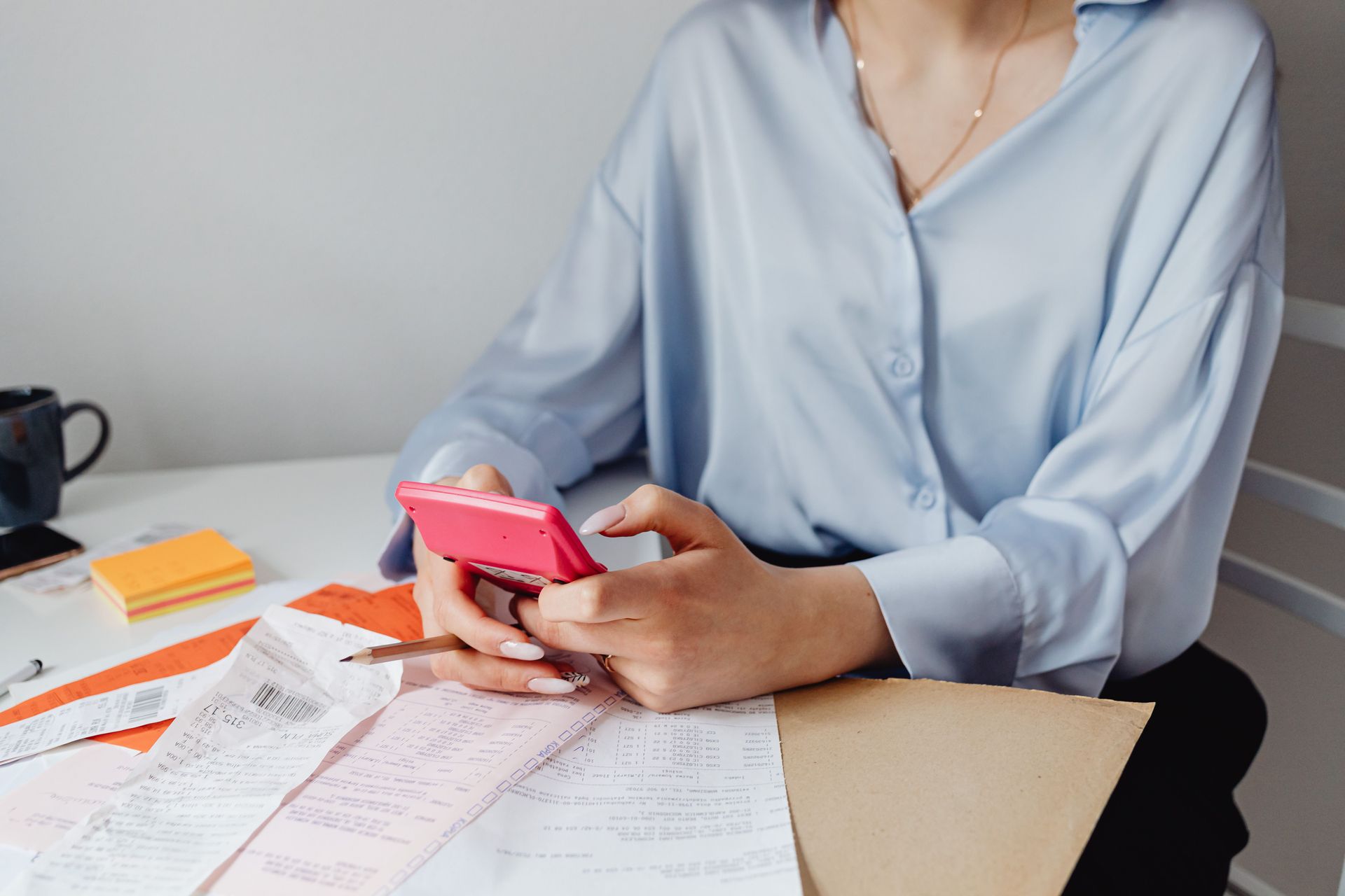 Person at a desk reviewing papers while holding a pink smartphone, with sticky notes nearby.