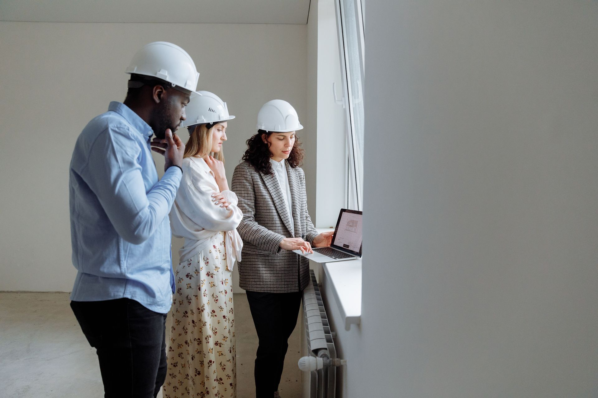 Three people in hard hats reviewing plans at a laptop in a bright unfinished room