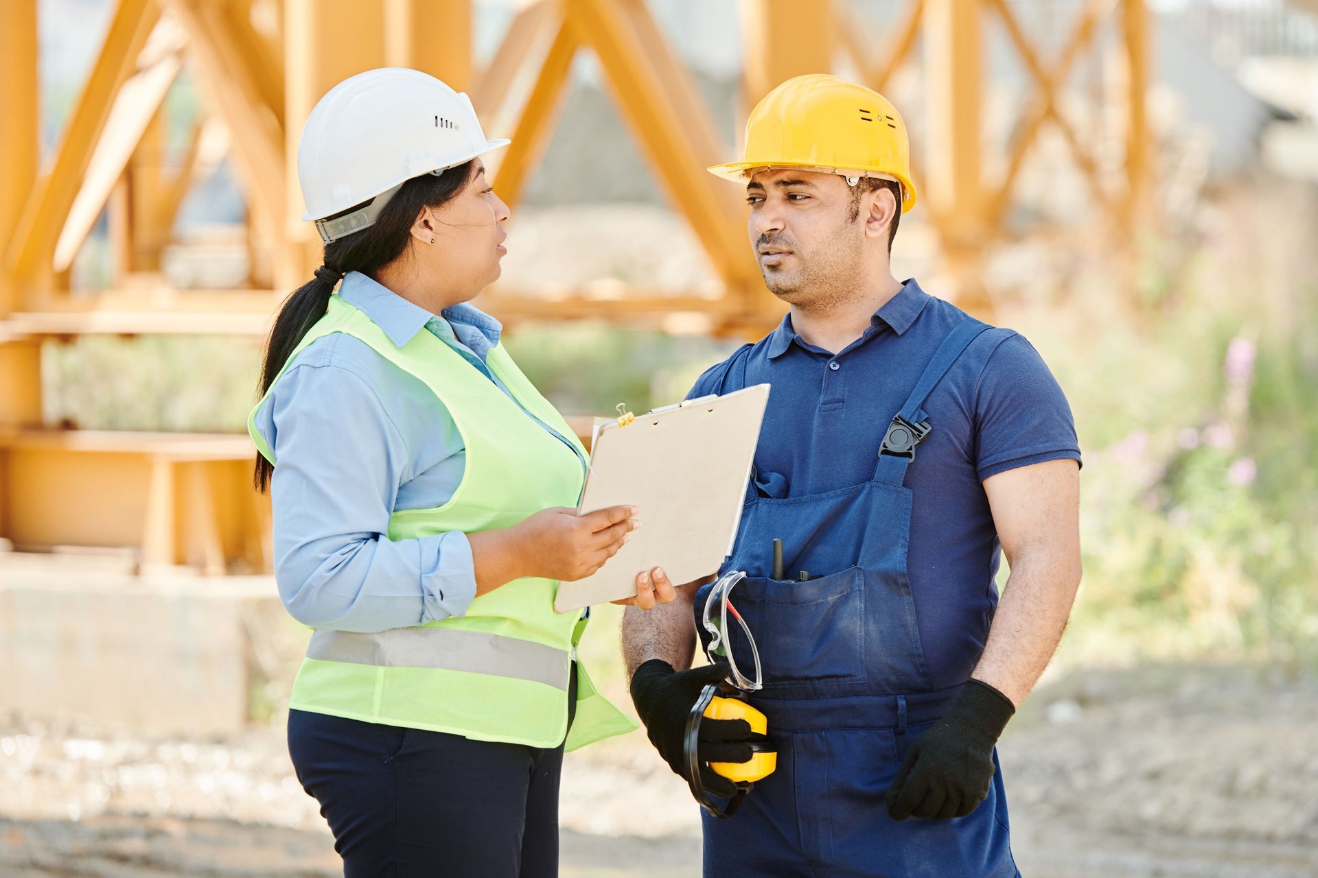 Two construction workers in hard hats discuss plans outdoors, one holding a clipboard and the other a tape measure.