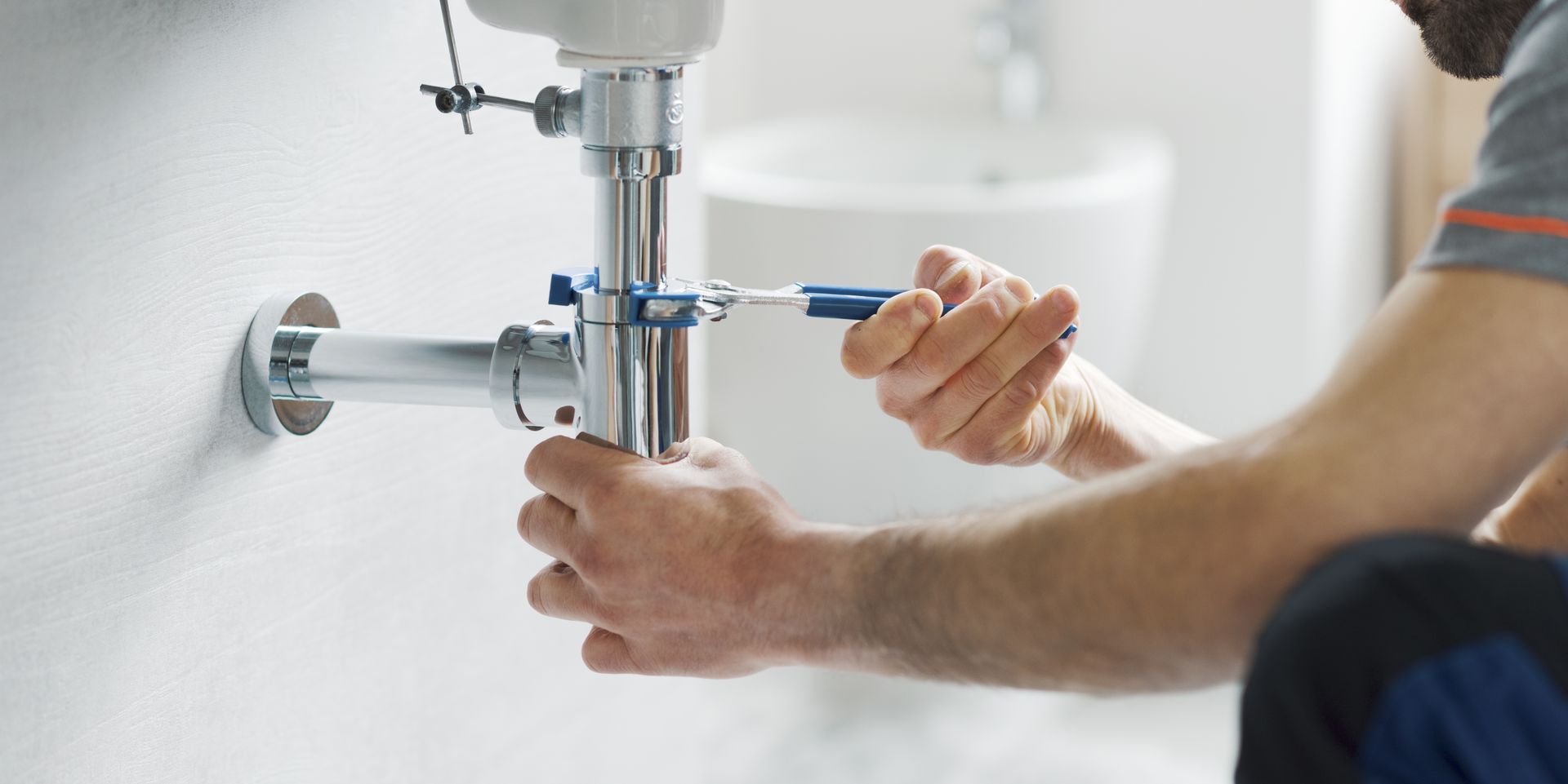 Person tightening a bathroom sink pipe with a wrench under a white basin