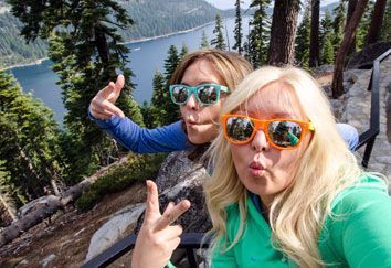 Two women wearing sunglasses are taking a selfie in front of a lake.