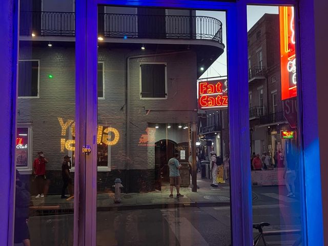 View through a window onto a New Orleans street at dusk, featuring brick buildings, neon signs, and people walking by.