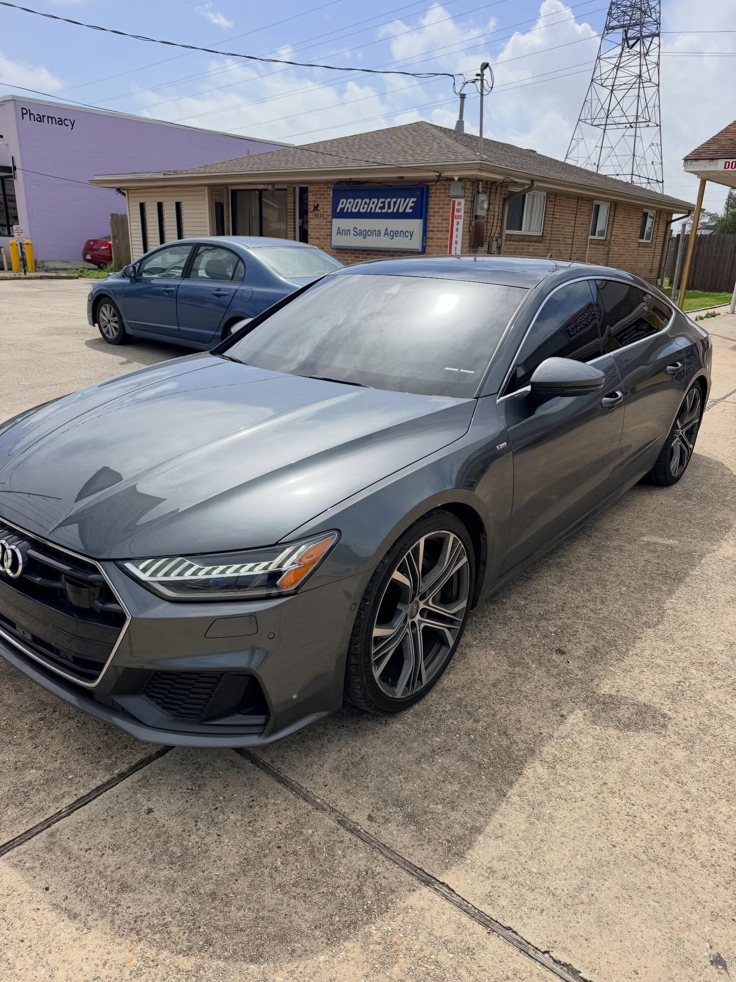 A gray Audi A7 parked on a concrete lot in front of a brick building on a sunny day.