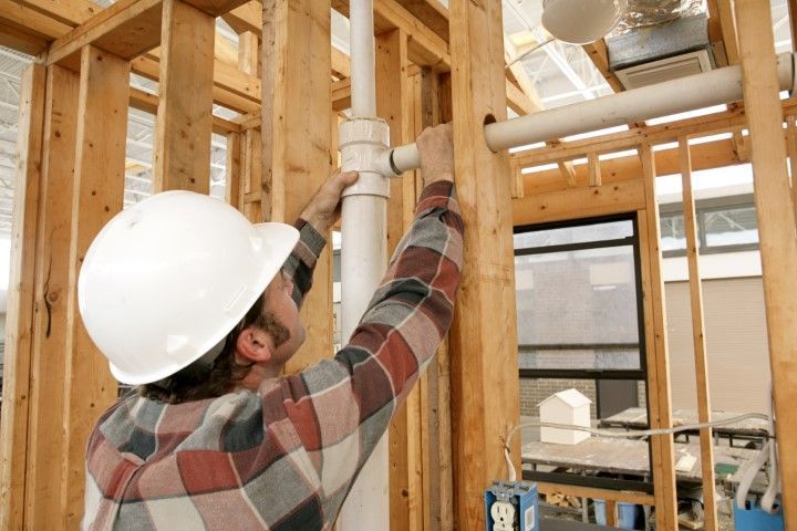 Construction worker in hard hat installing pipes inside a wooden-framed building.