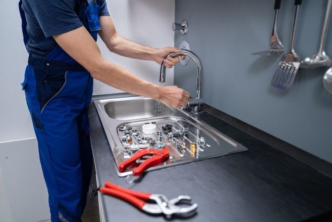 Person in blue workwear washing dishes at a stainless-steel sink in a kitchen, with utensils on the counter.
