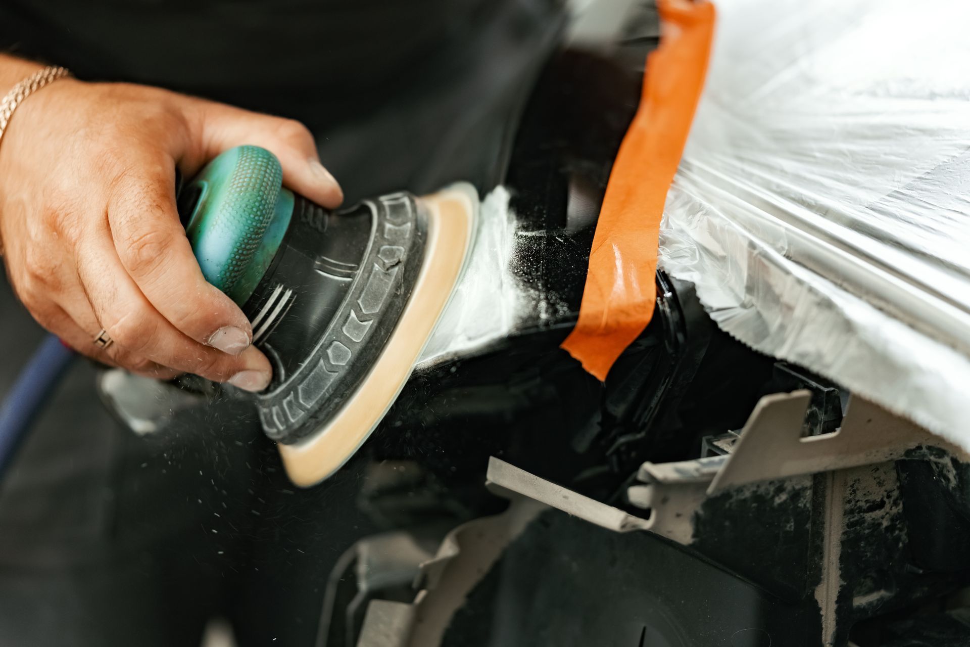 Hand using a green buffer to sand a car panel with orange tape masking the edge