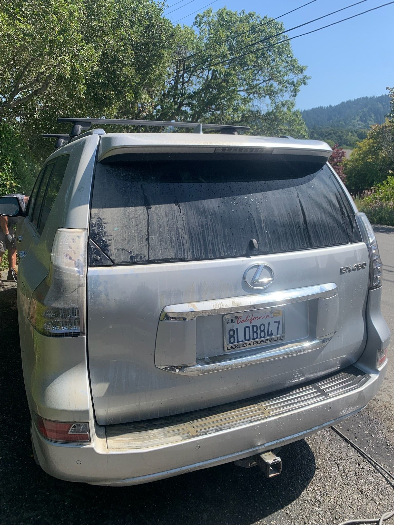 Rear view of a silver SUV parked on a gravel roadside, with trees and mountains in the background.