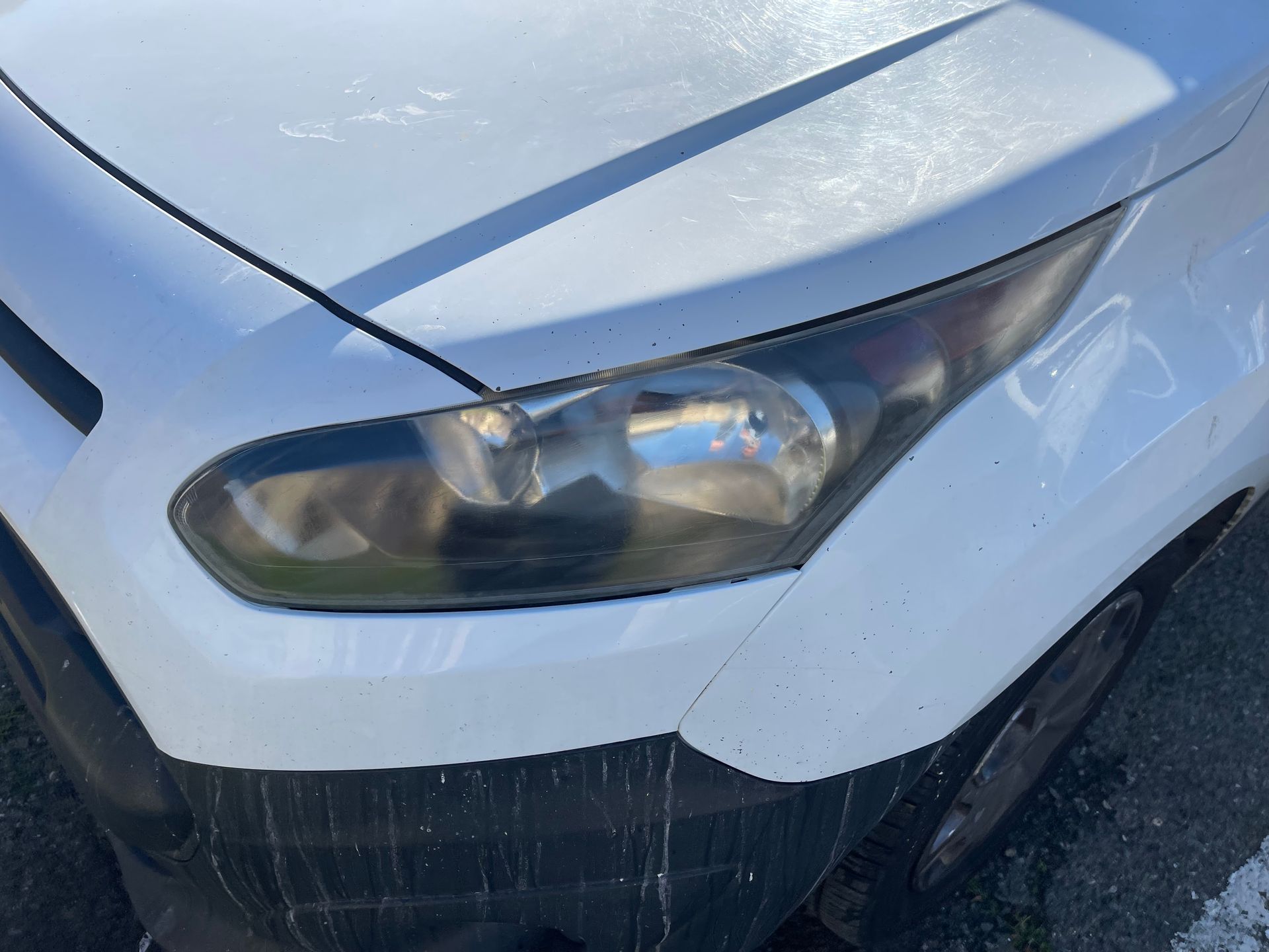 Front corner of a white car with a damaged headlight and scrape marks on the bumper