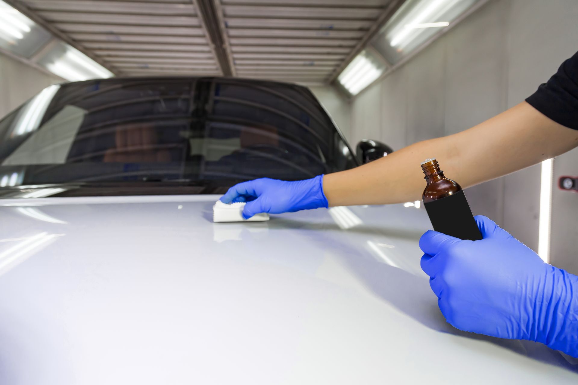Gloved hands polishing a white car hood in a garage, with a bottle of detailing liquid nearby