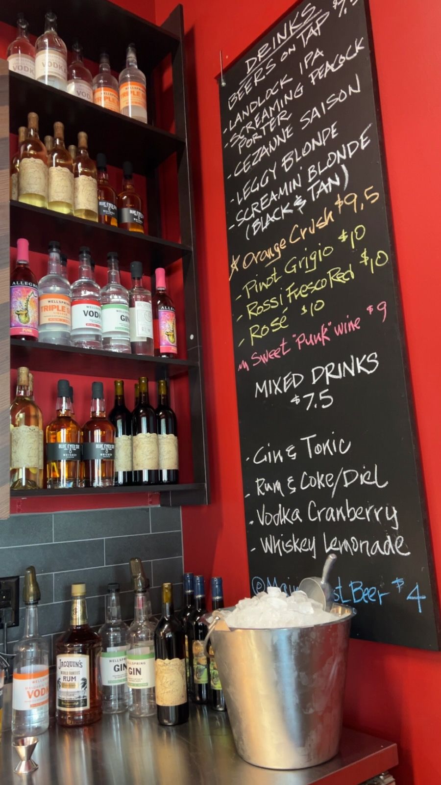 A bar area featuring a chalkboard menu on a bright red wall, a stainless steel counter, shelves of liquor, and beer taps.