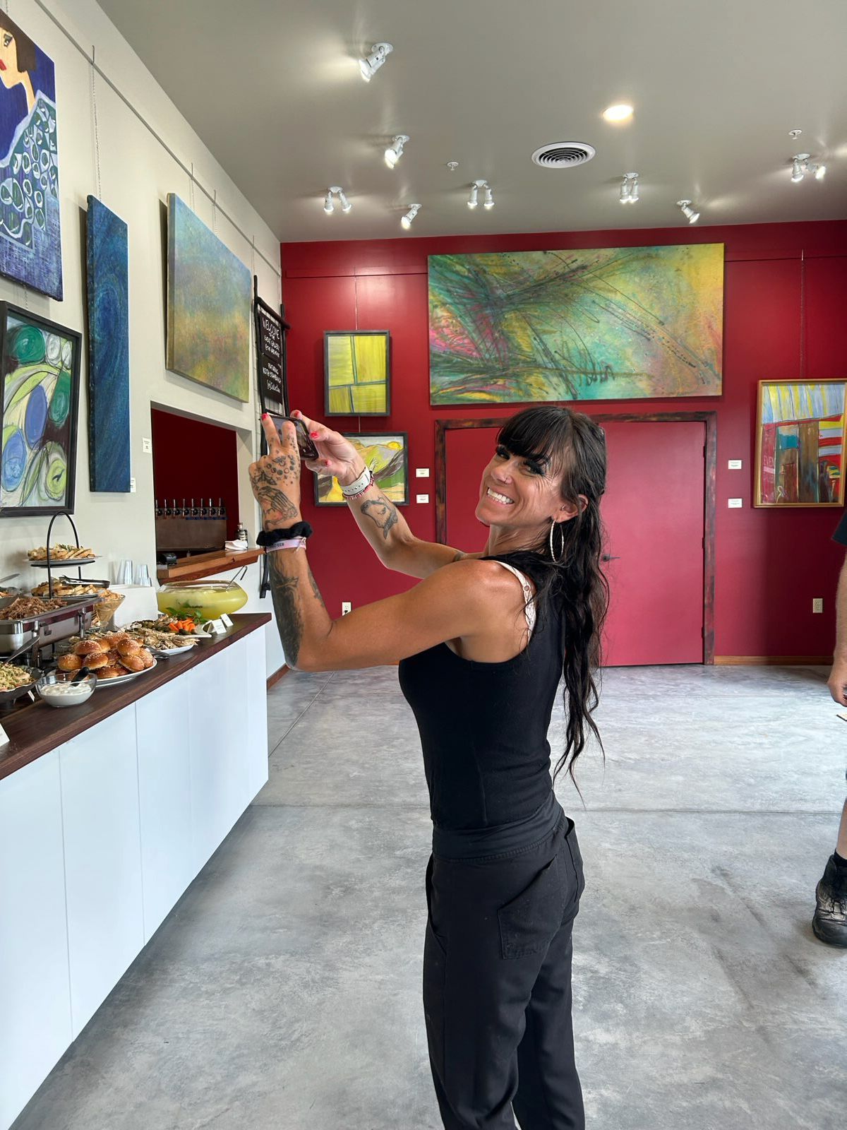 An art gallery with concrete floors, a red accent wall, large windows, and framed paintings displayed on white walls.