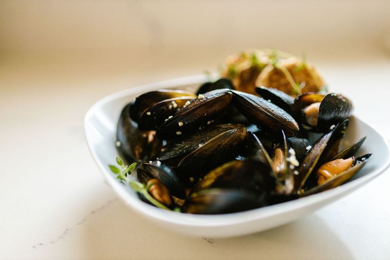A bowl of steamed clams in a light broth, garnished with a lemon slice and herbs, served with chopsticks on a wooden table.