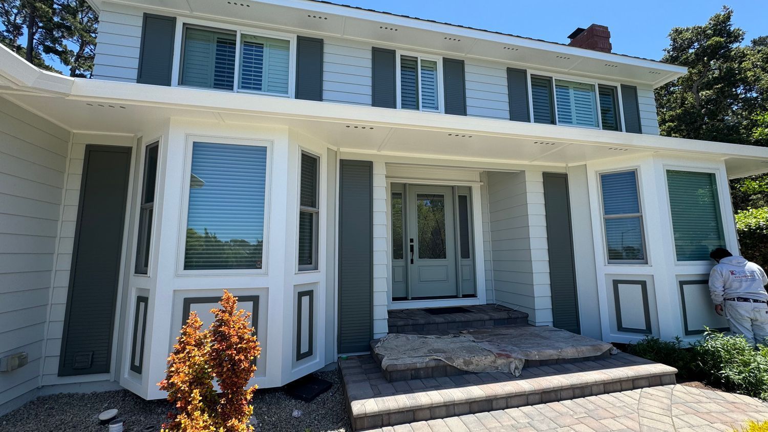 Two-story house with light blue siding, white trim, and gray shutters. A person paints on the side.