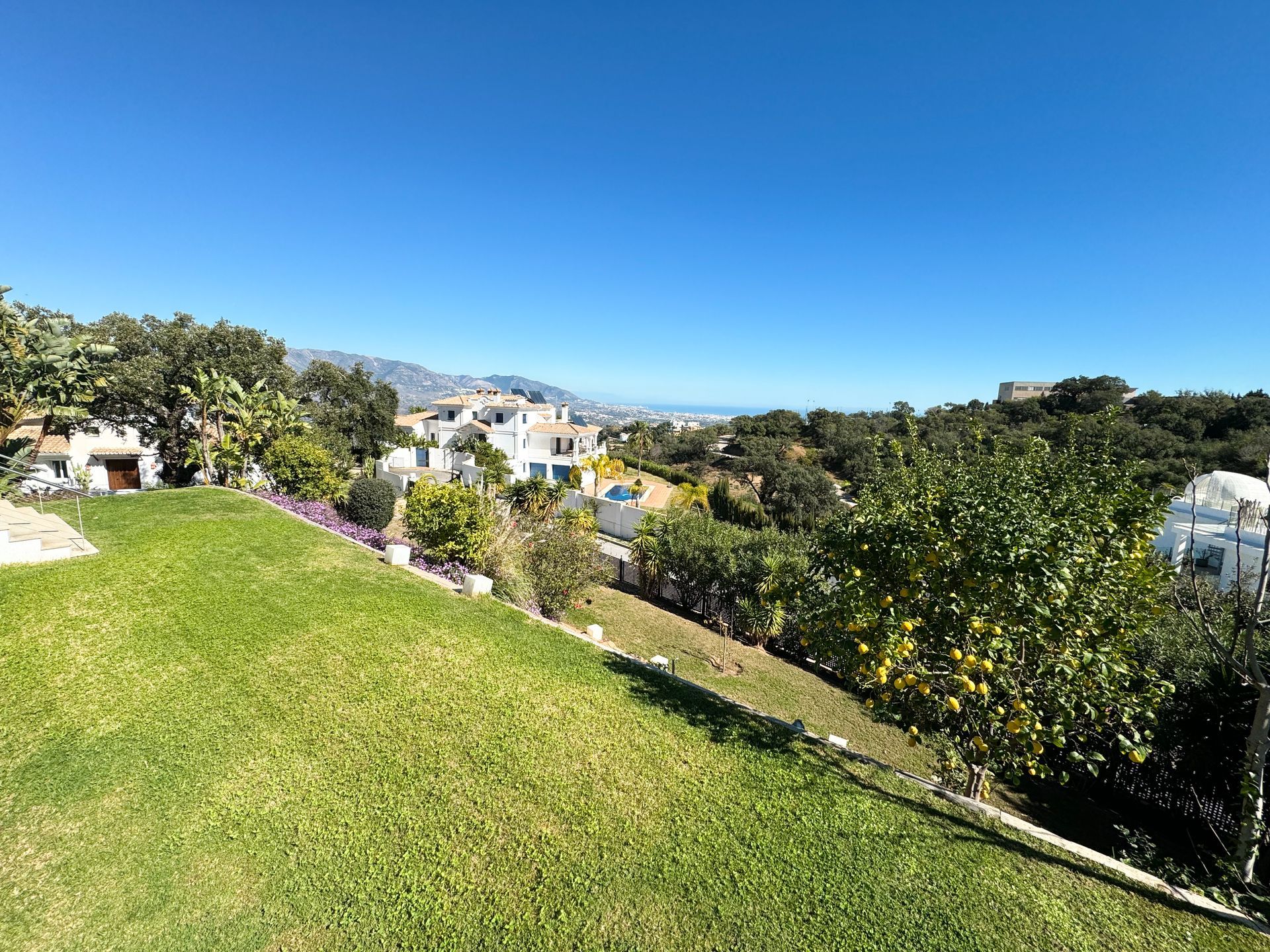 Lush green lawn slopes down to a view of white villas, trees, and a distant blue sea under a clear sky.