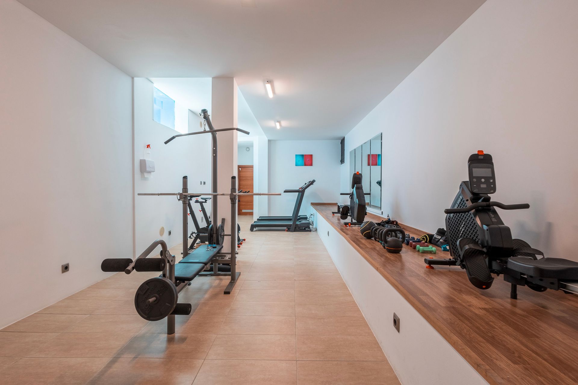 Gym interior with weight bench, pull-up bar, treadmill, rowing machine, and free weights. White walls and a wooden counter.