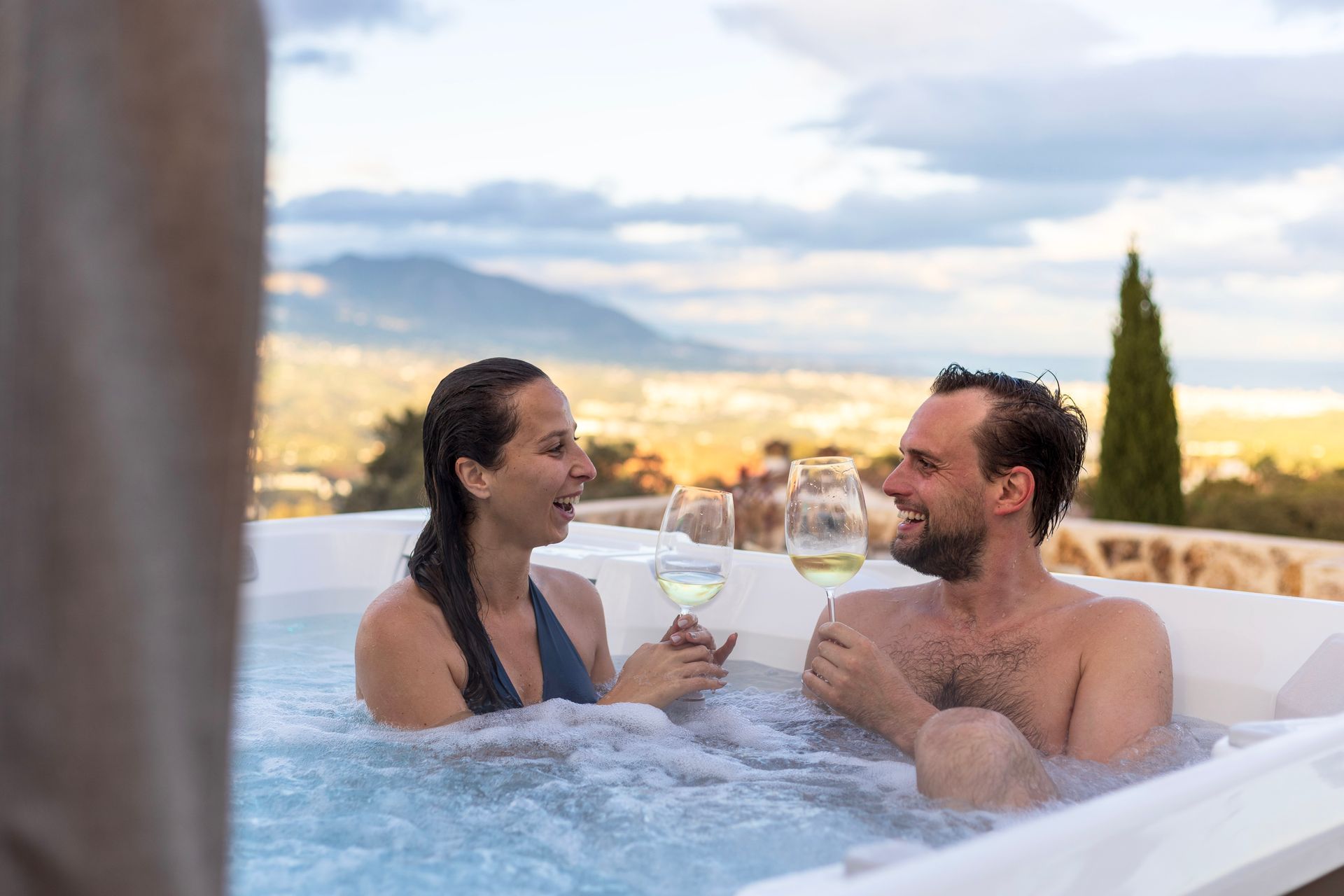 A couple in a hot tub, smiling and holding glasses of wine, with a scenic mountain view in the background.