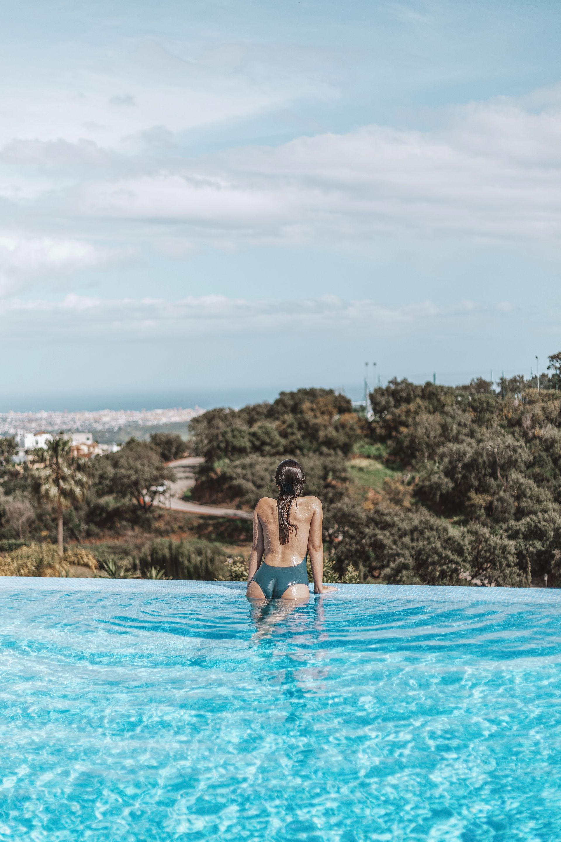 Woman in a blue swimsuit entering an infinity pool overlooking a green hillside and city skyline on a partly cloudy day.
