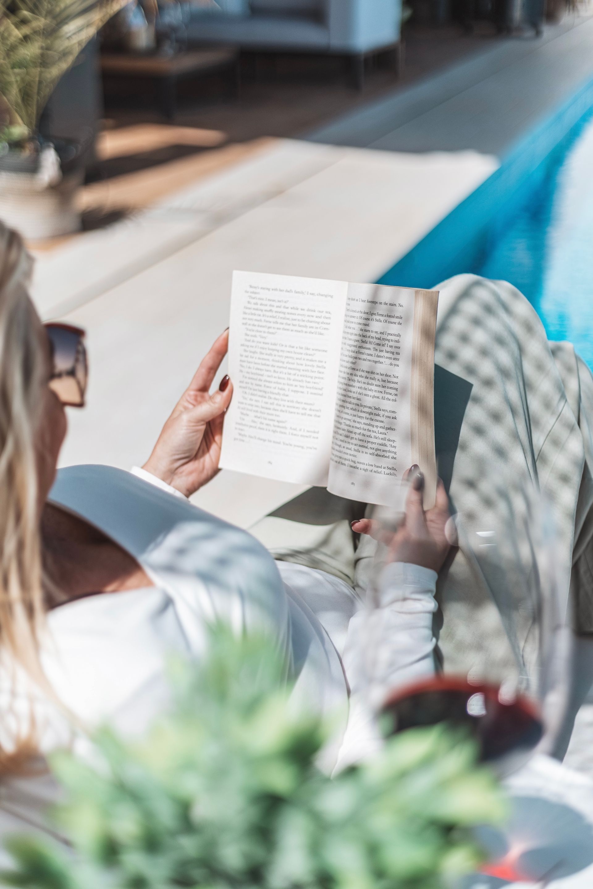 Woman wearing sunglasses reading a book near a pool on a sunny day.