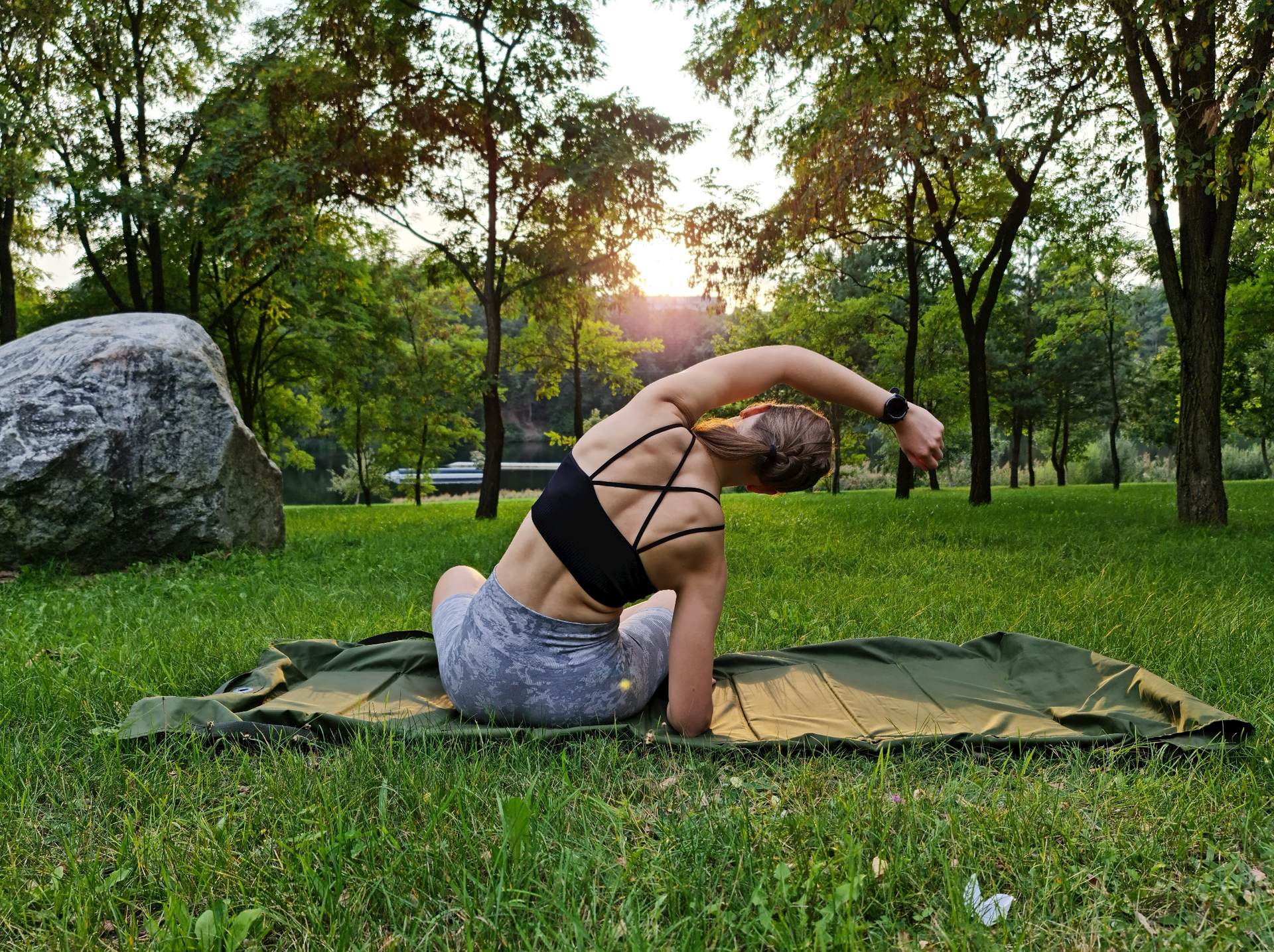 Woman in a black sports bra stretches on a mat in a grassy park with trees and the sun setting in the background.