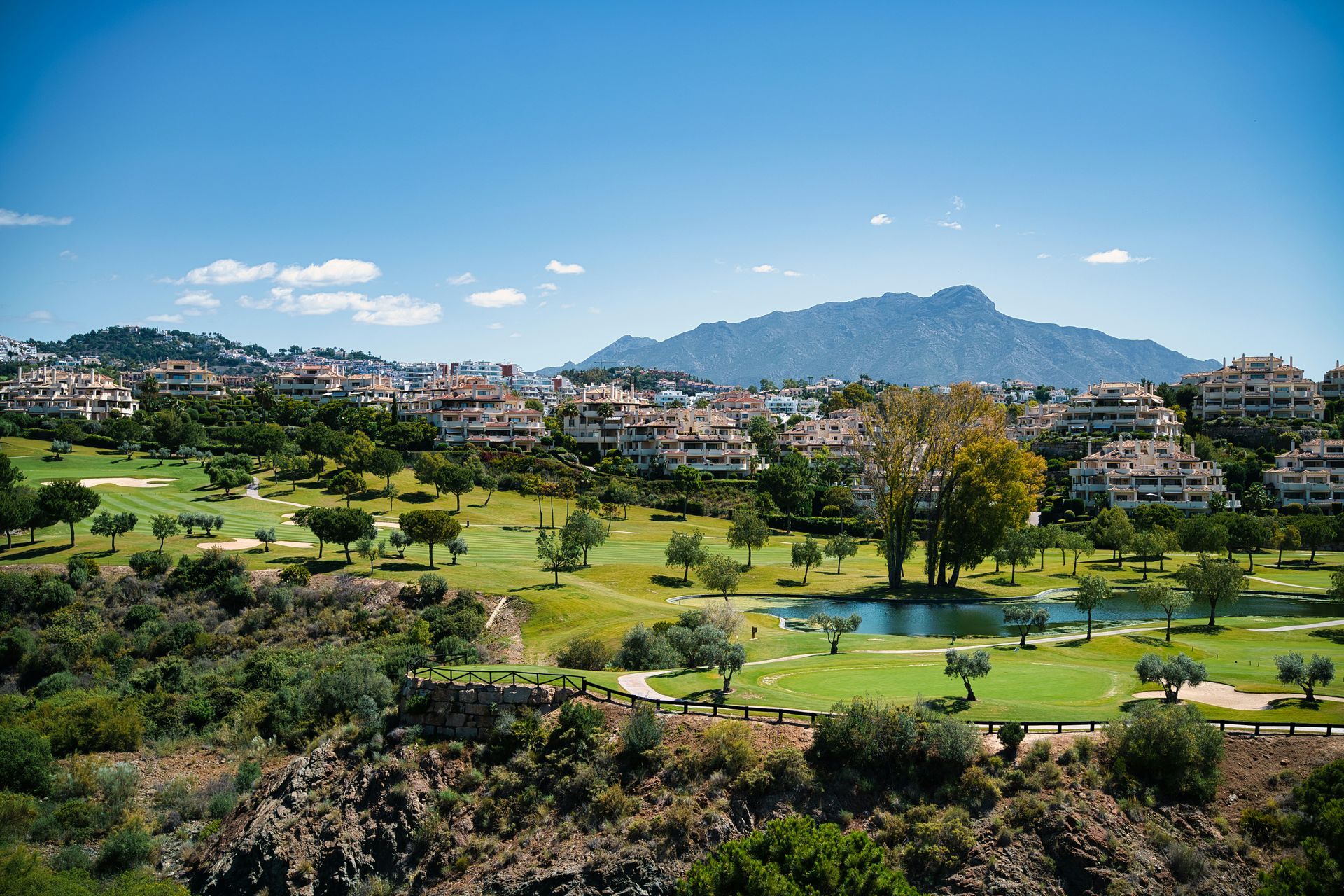 A sunny golf course with townhouses, trees, and a mountain backdrop under a clear blue sky.