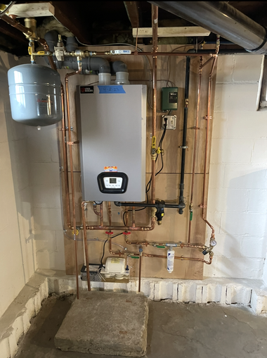 A modern grey tankless water heater with copper pipes and a beige expansion tank mounted on a white wall.