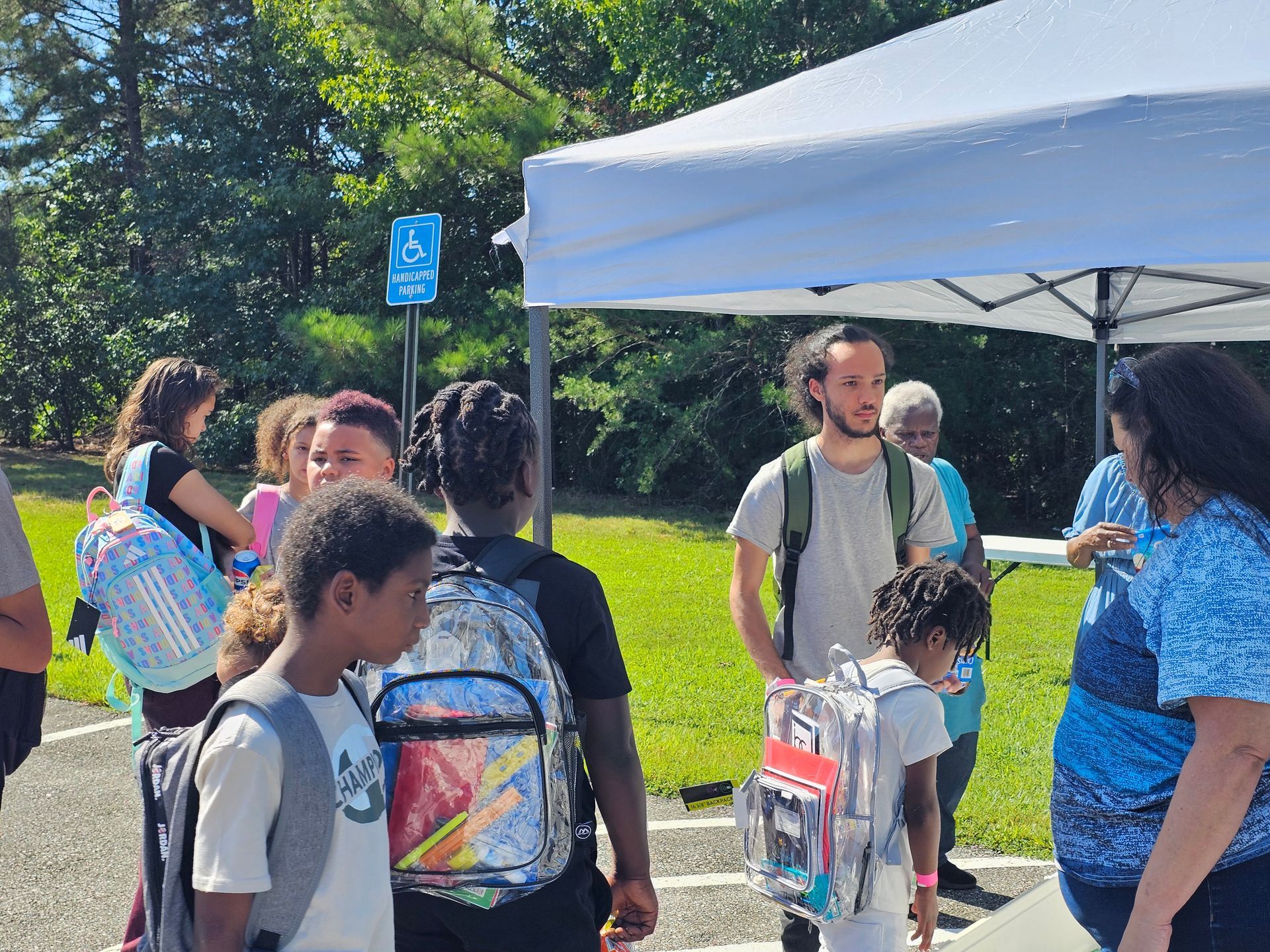 Children gathered under a white canopy tent at an outdoor event, with backpacks and trees in the background.
