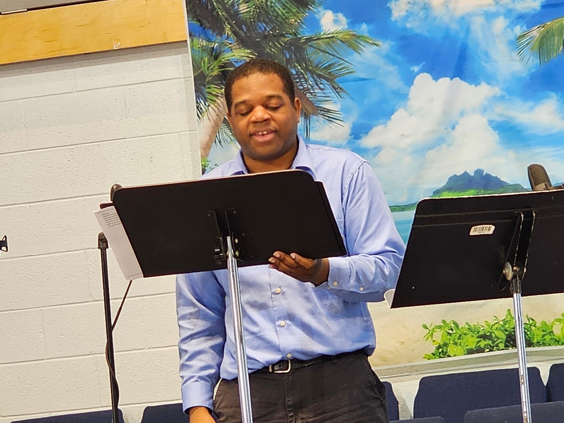 Man speaking at a microphone behind a music stand, with a painted sky mural in the background