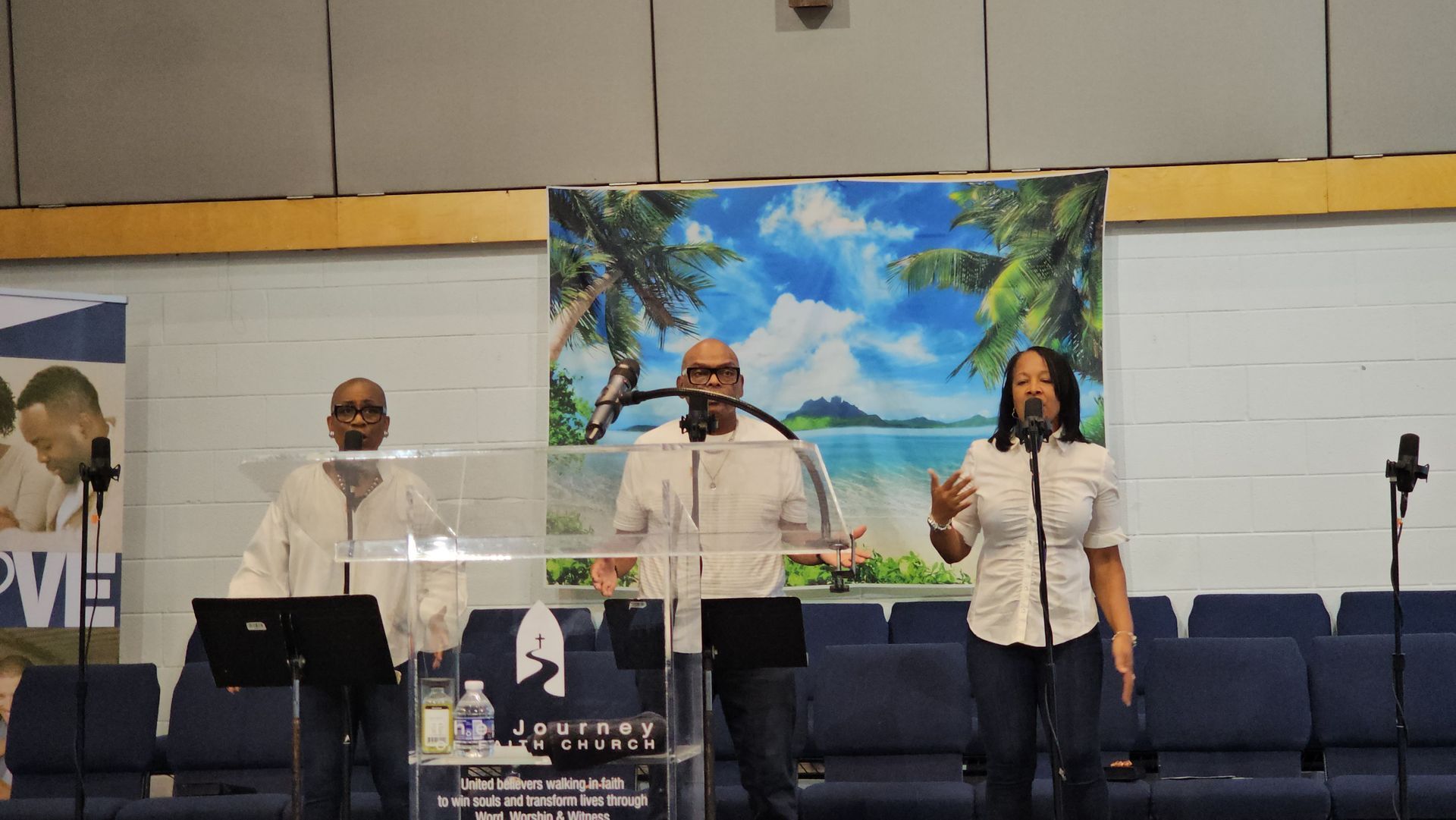 Three singers perform on a church stage with microphones, a blue backdrop, and a painting behind them.