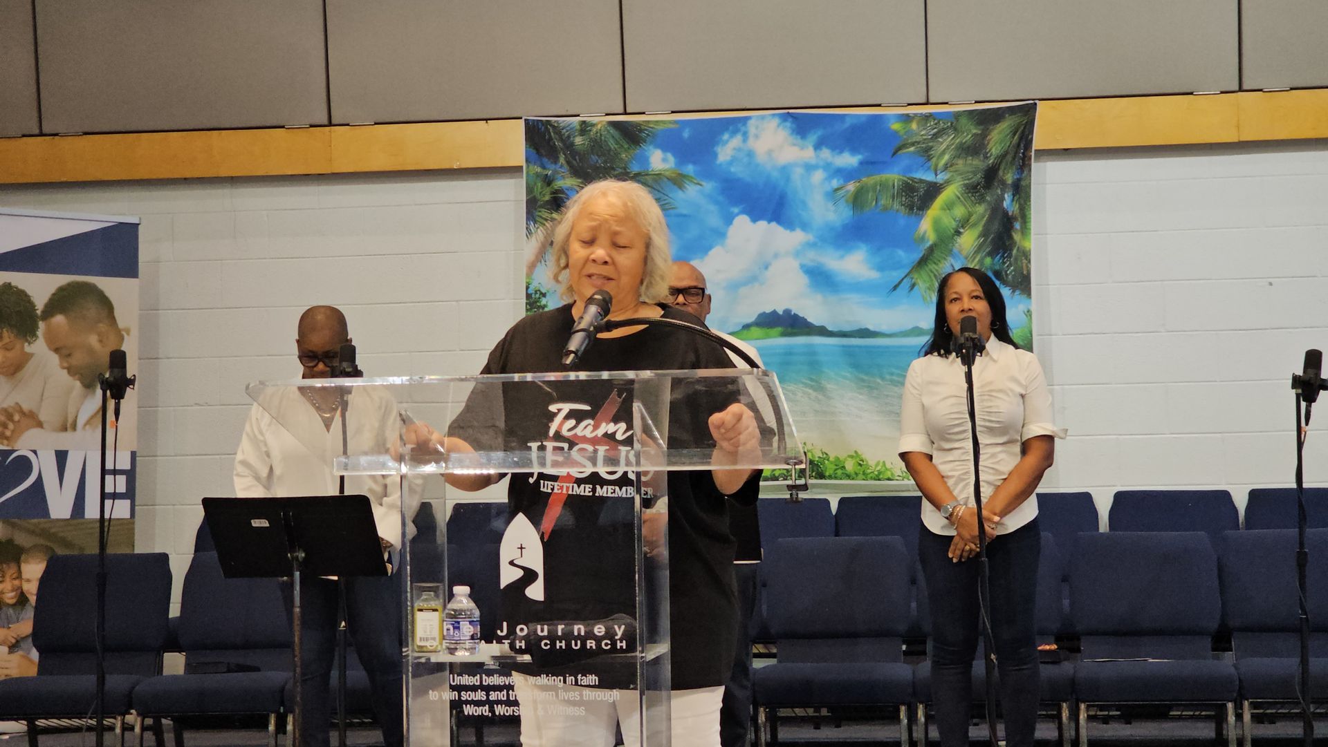Woman speaking at clear podium in church, with two people standing behind her and blue chairs in the background