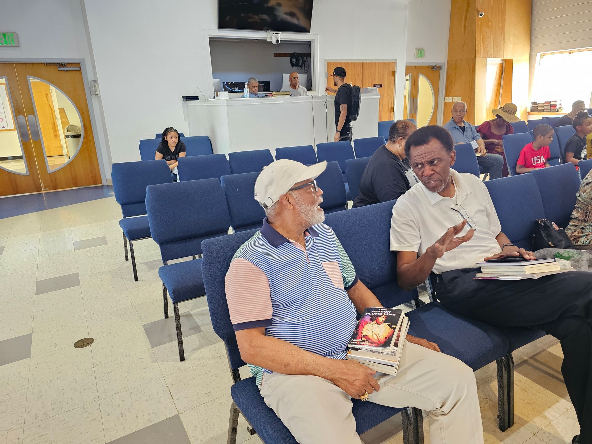 People seated in a waiting room, talking and holding papers, with rows of blue chairs and a TV in the background.