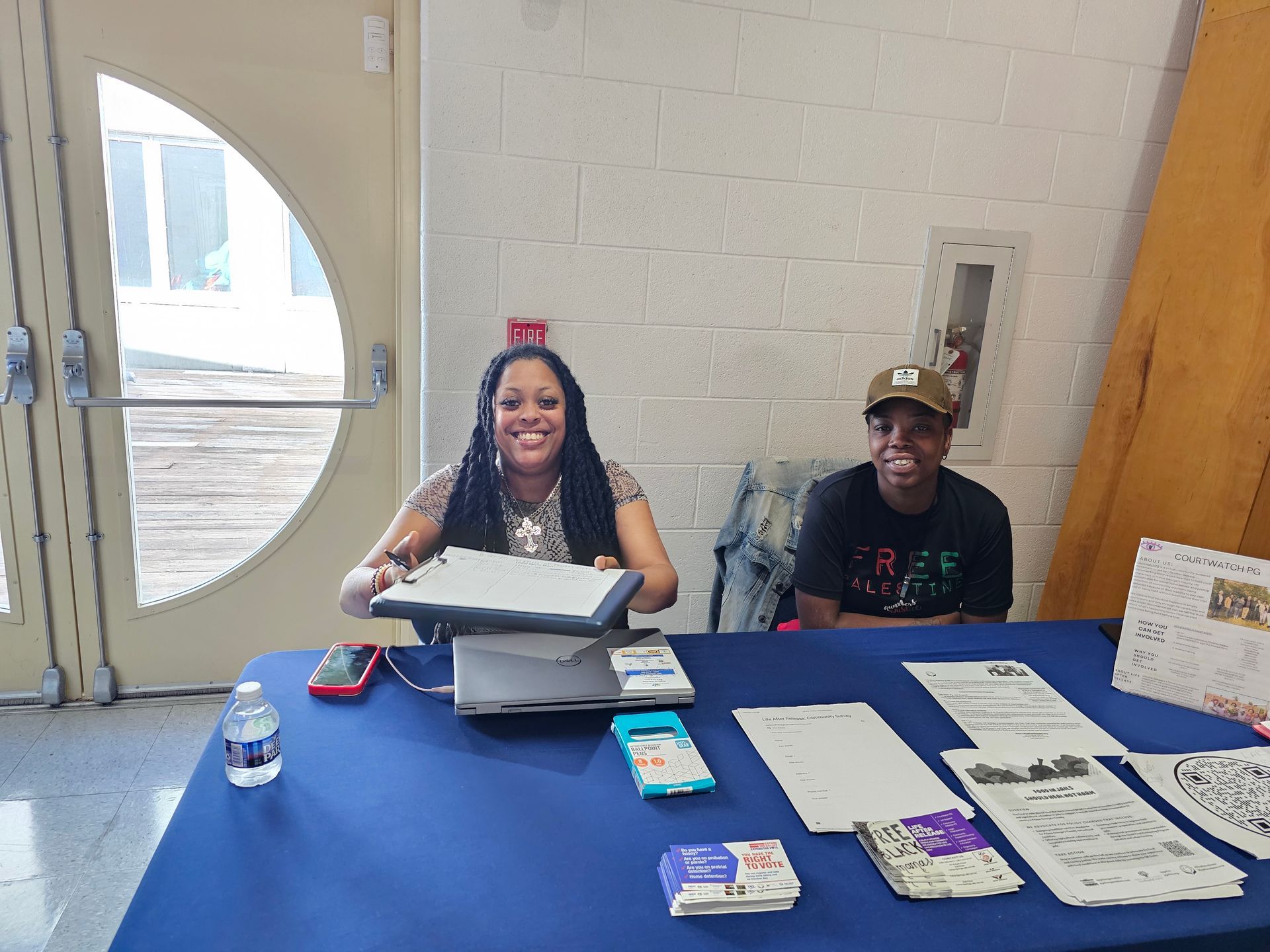 Two people seated at a blue table with papers, forms, and small items in a lobby near a glass door