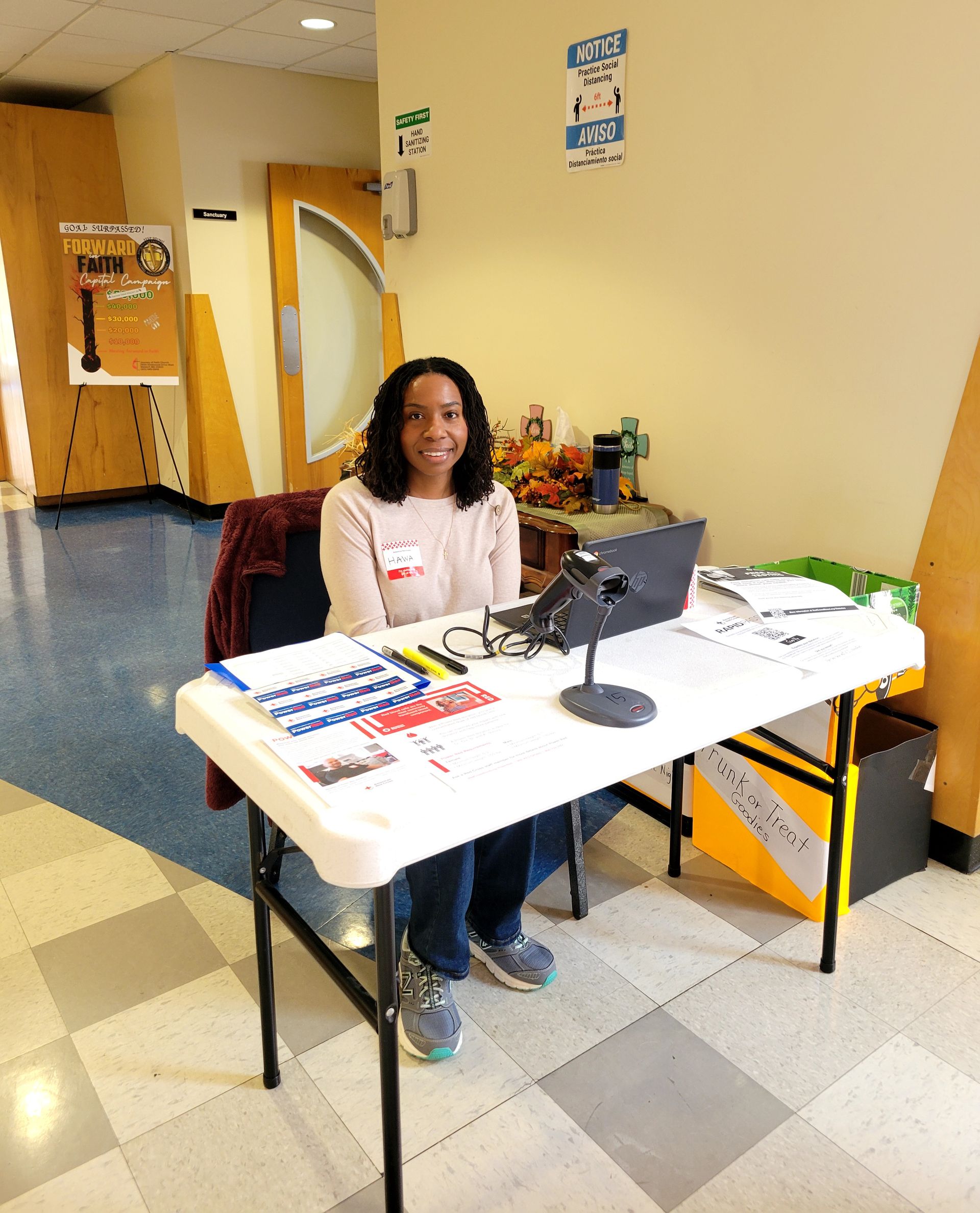 Person seated at a table in a hallway, with papers, a laptop, and event materials.