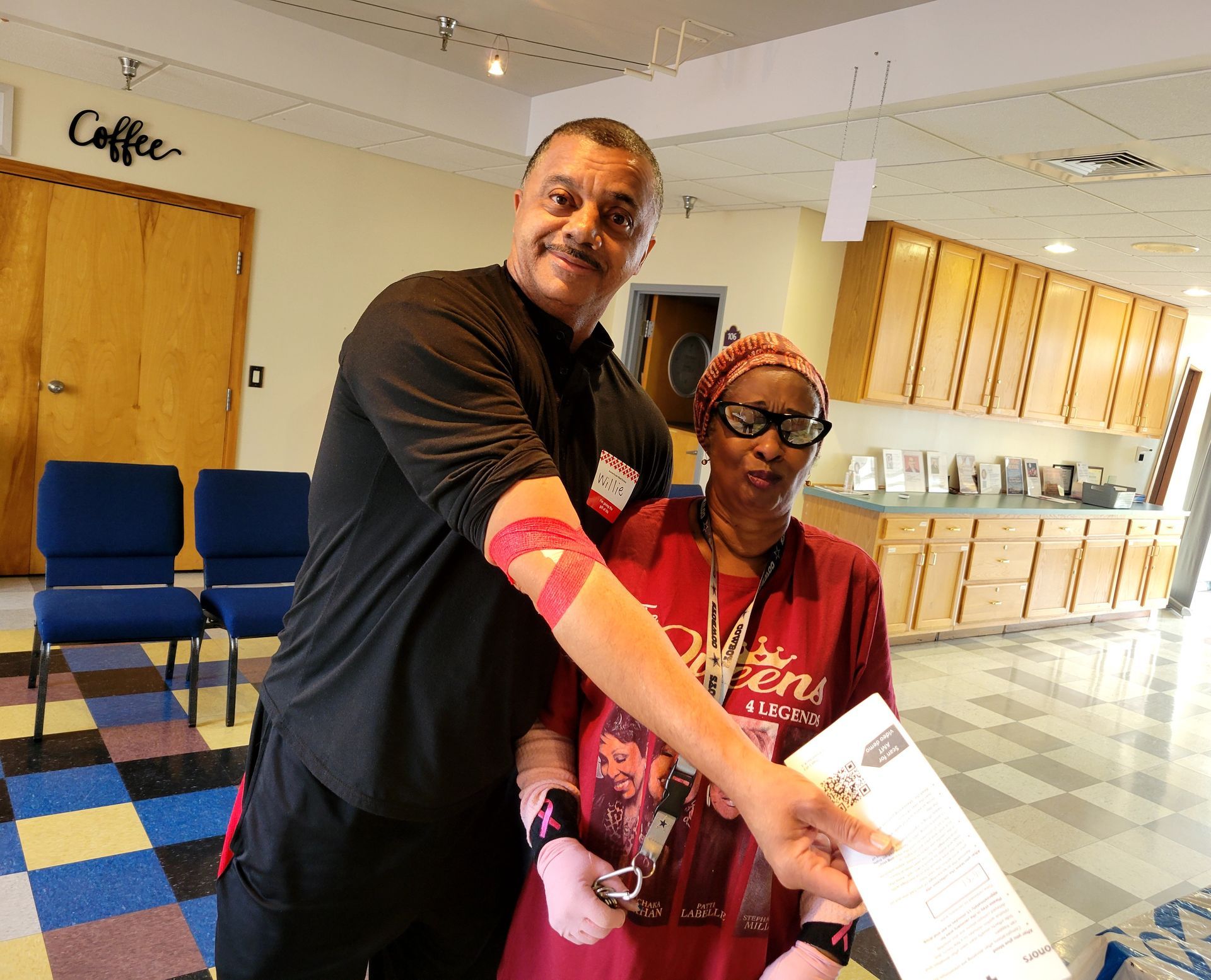 Two people smiling in a hallway, one pointing at a sheet of paper near a sign for the coffee area.
