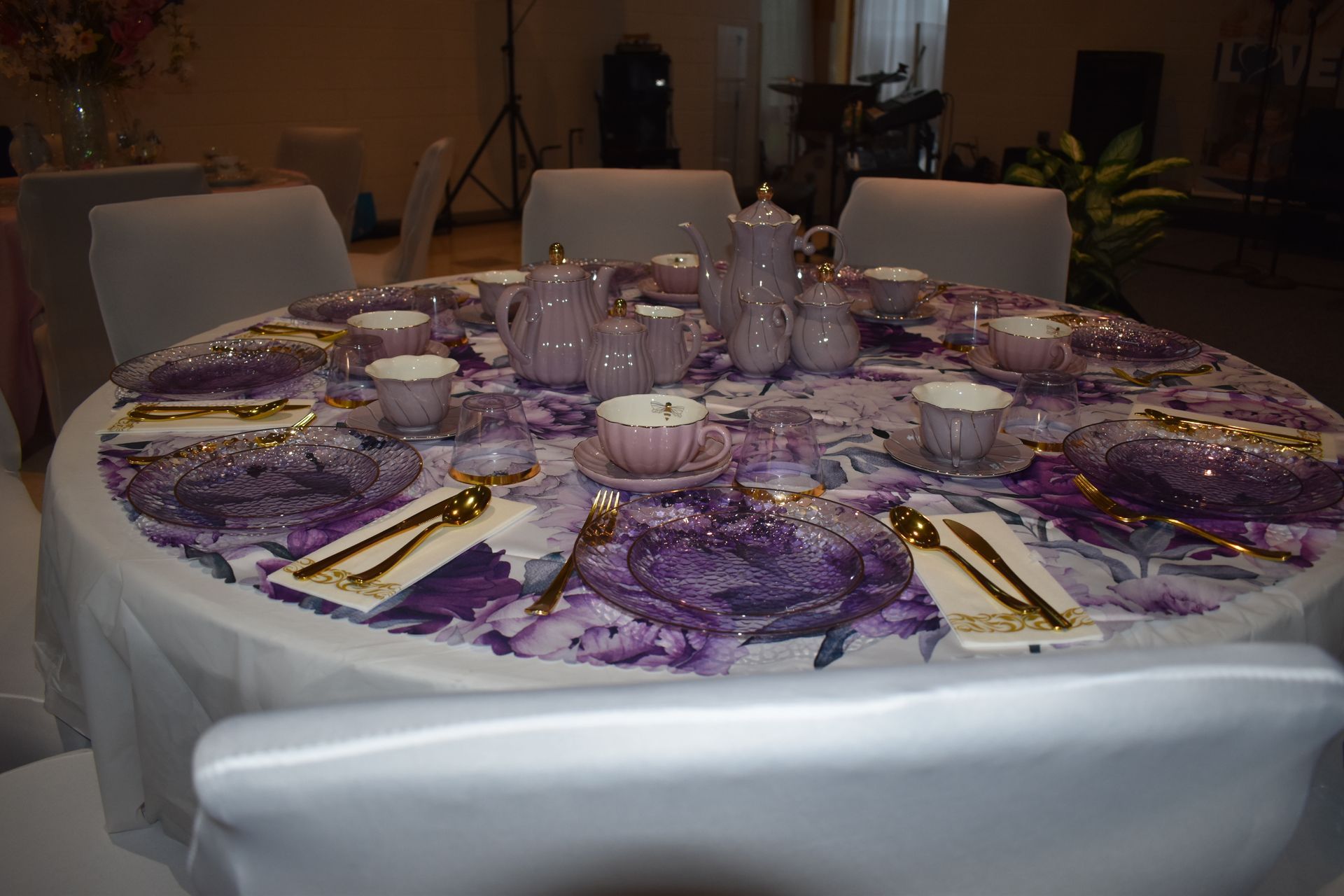 Round dining table set with purple floral tablecloth, white china, and white chairs in a dimly lit room