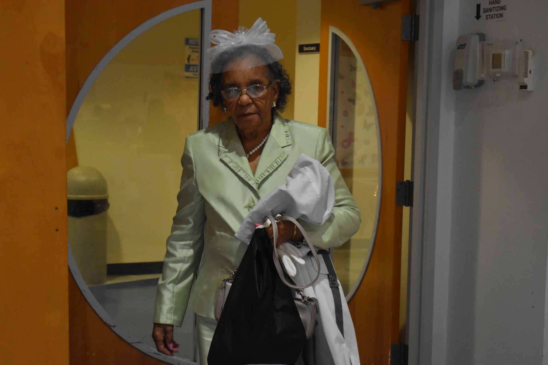 Elderly woman in a light green coat stands in a doorway, holding a white jacket and black bag.