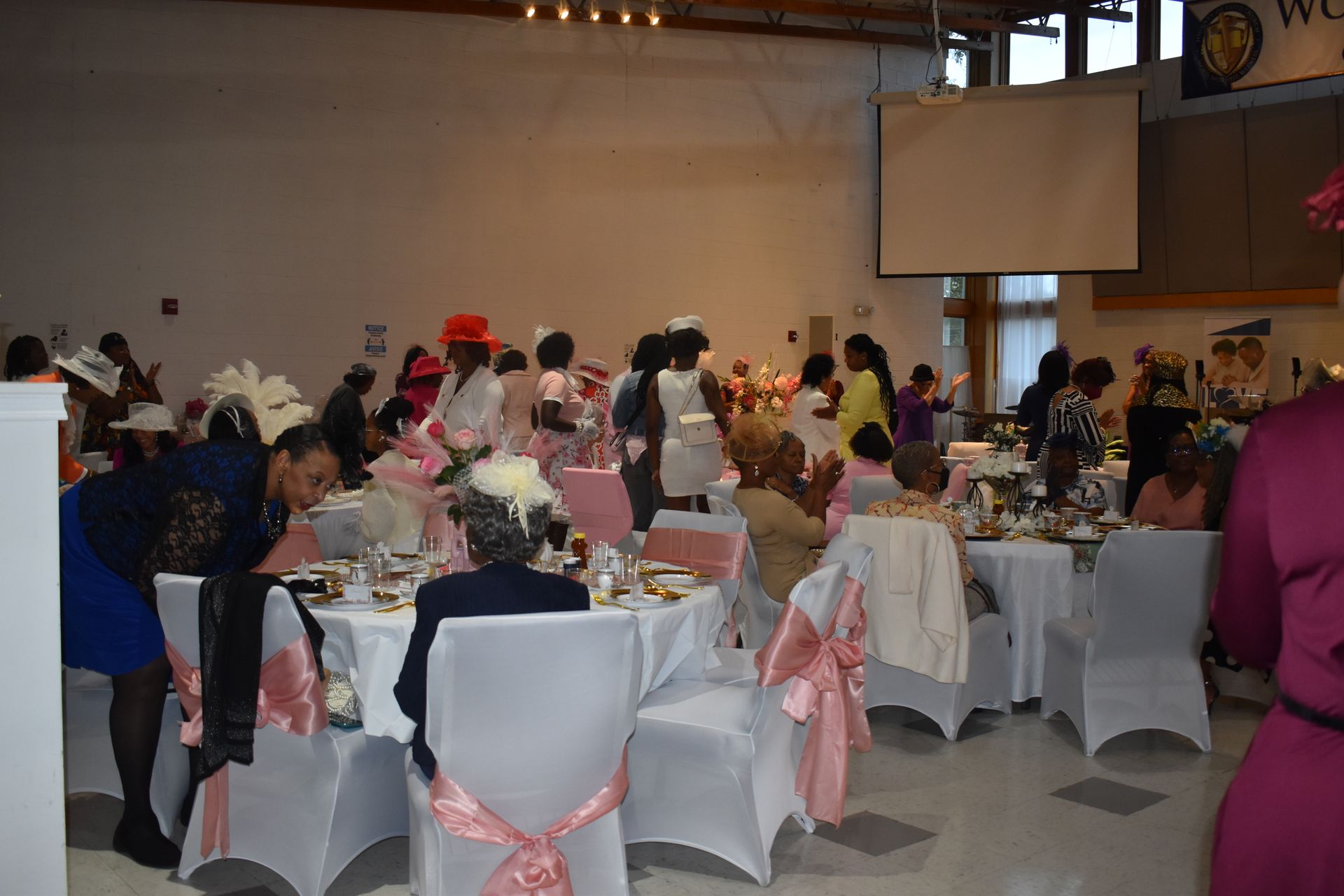 Decorated banquet hall with many guests seated at tables, white covers and pink ribbons on chairs