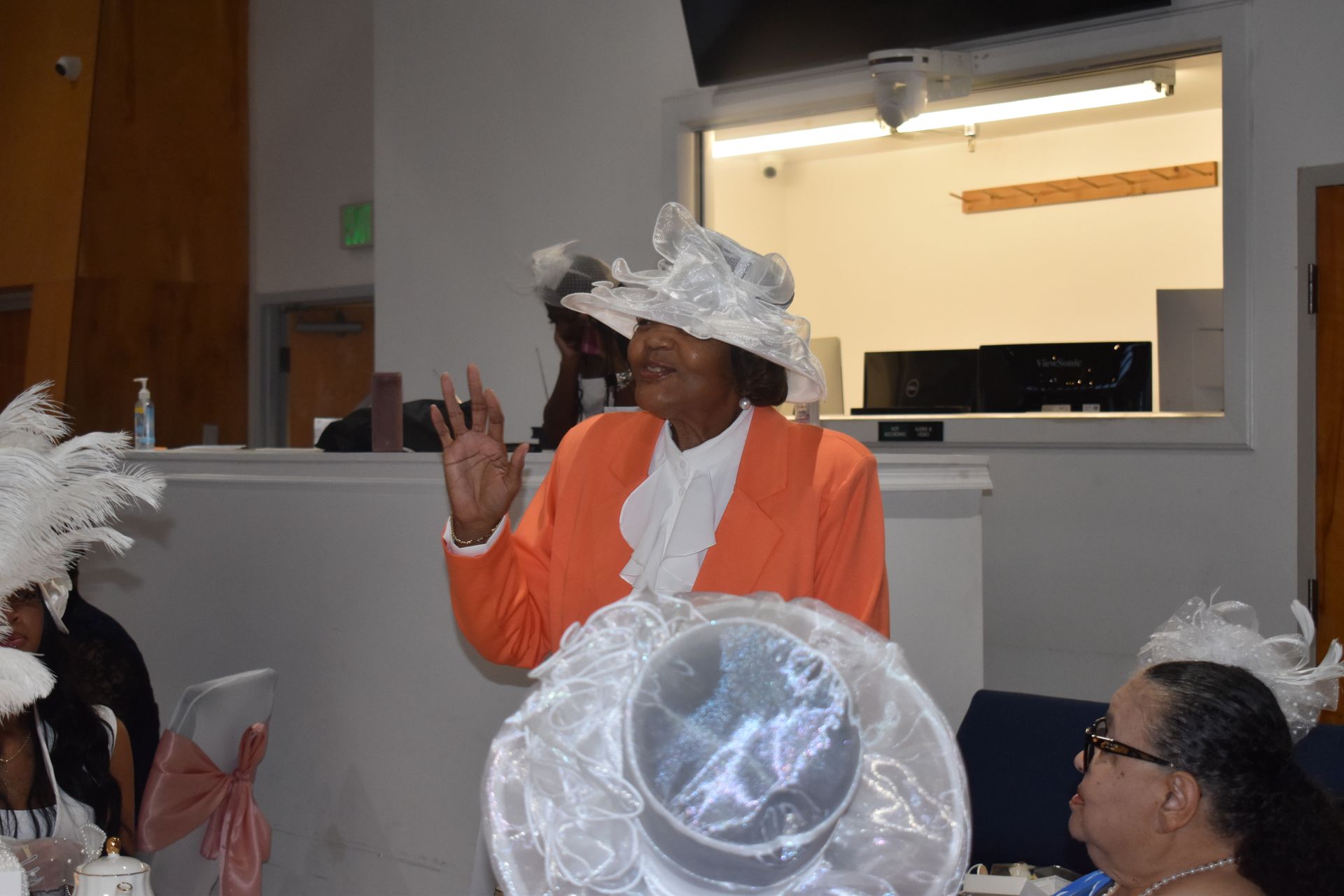 Person in orange jacket and white hat speaking indoors at a gathering