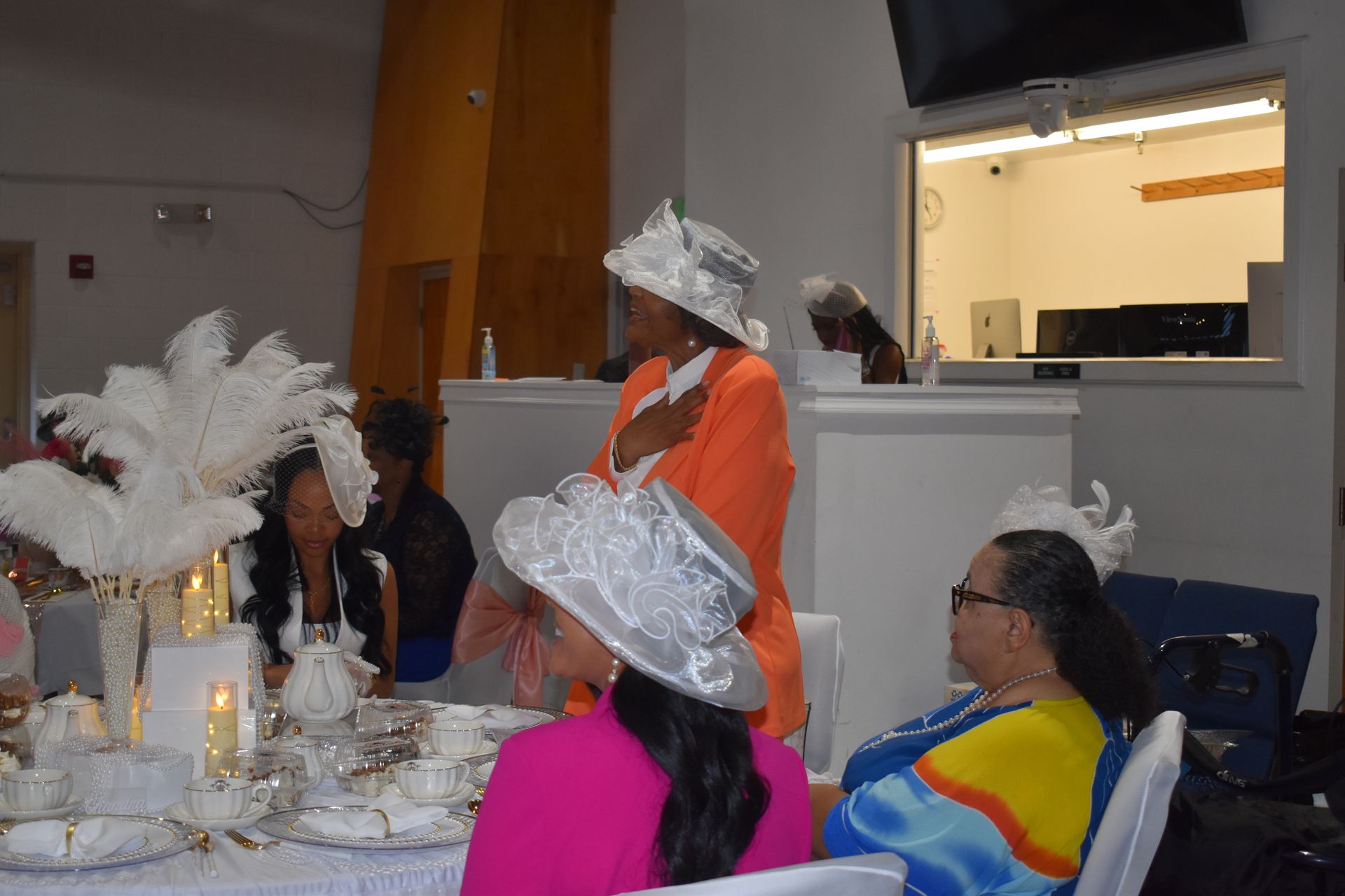 People seated at a decorated banquet table watching a woman in an orange outfit and silver hat speak