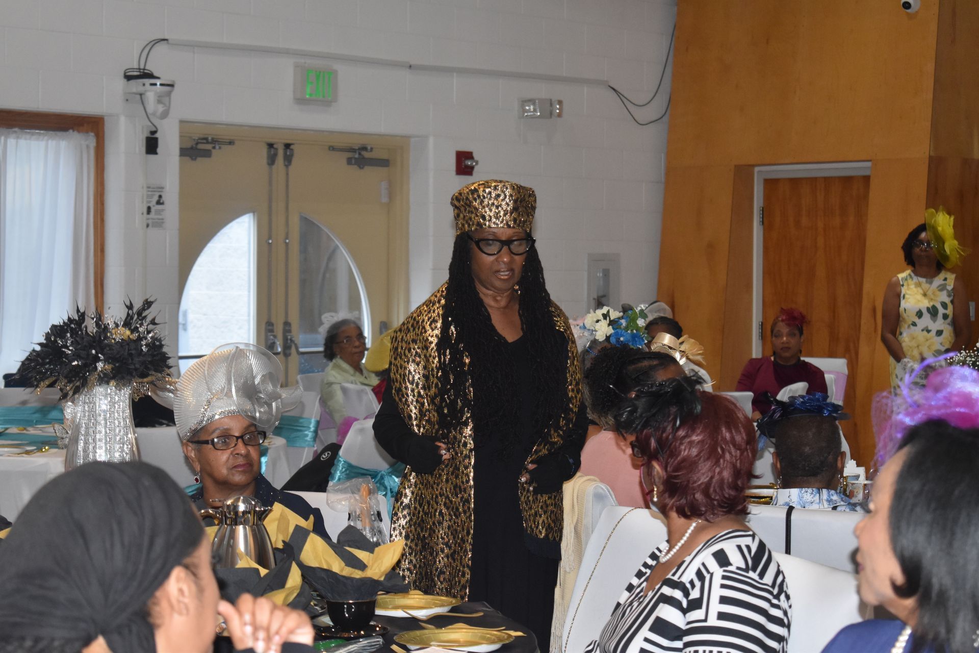 Woman in gold headdress stands among seated guests at a banquet in a decorated hall
