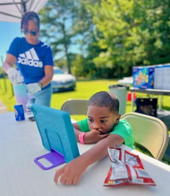 Child at an outdoor table using a tablet, with an adult serving food in the background.