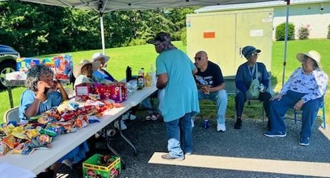 People gathered under a canopy at a community picnic, eating and chatting beside a table of food.