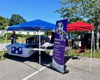 Outdoor community booth with blue and red tents, table displays, and a purple informational banner in a parking lot
