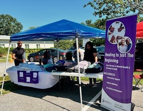 Outdoor outreach booth under a blue canopy with a purple banner and table at a parking lot event