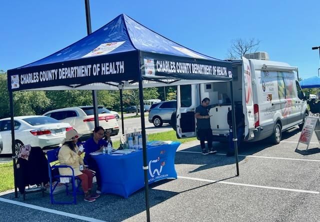 Public health outreach tent in a parking lot, with staff seated at a blue table beside a mobile clinic van.