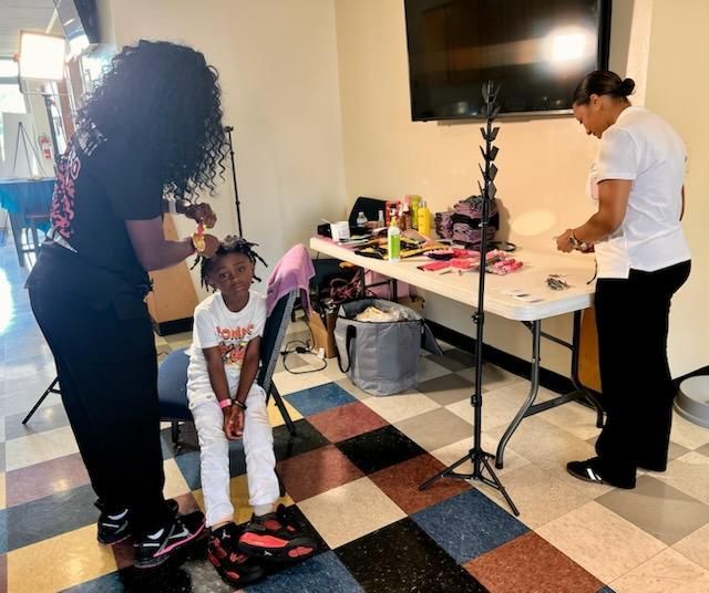 Two people styling a child’s hair in a living room with a table of supplies and a TV on the wall