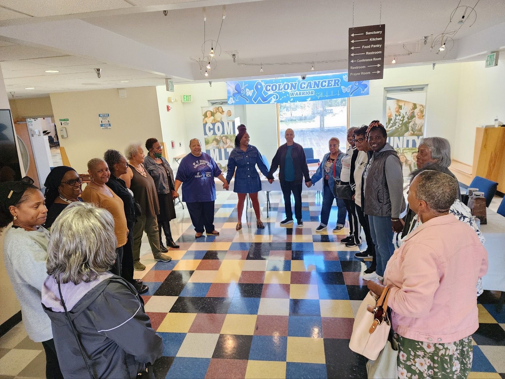People holding hands in a circle on a colorful checkered floor in a bright indoor room.