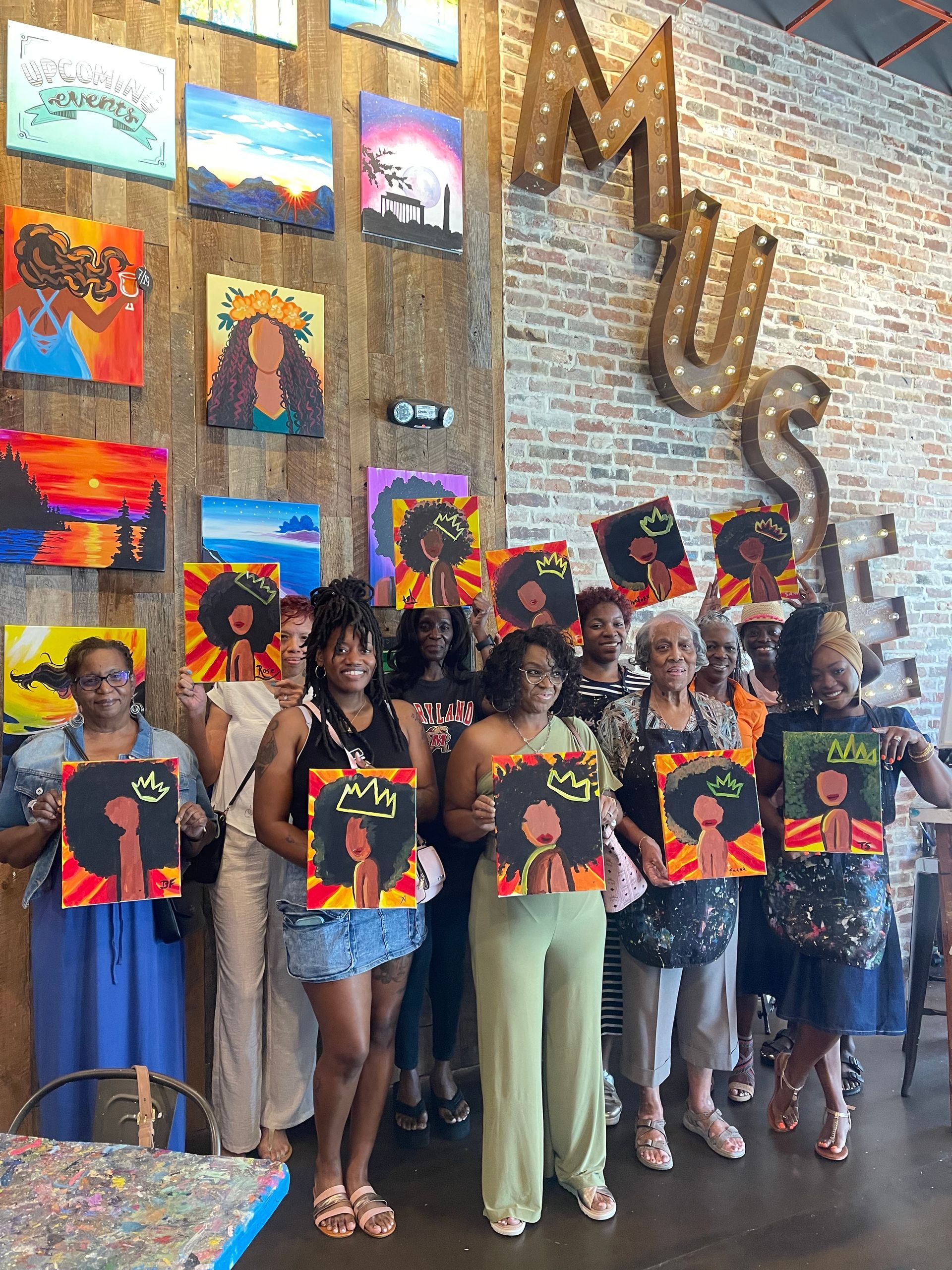 Group holding colorful paintings in front of a mural wall with large “MUS” letters