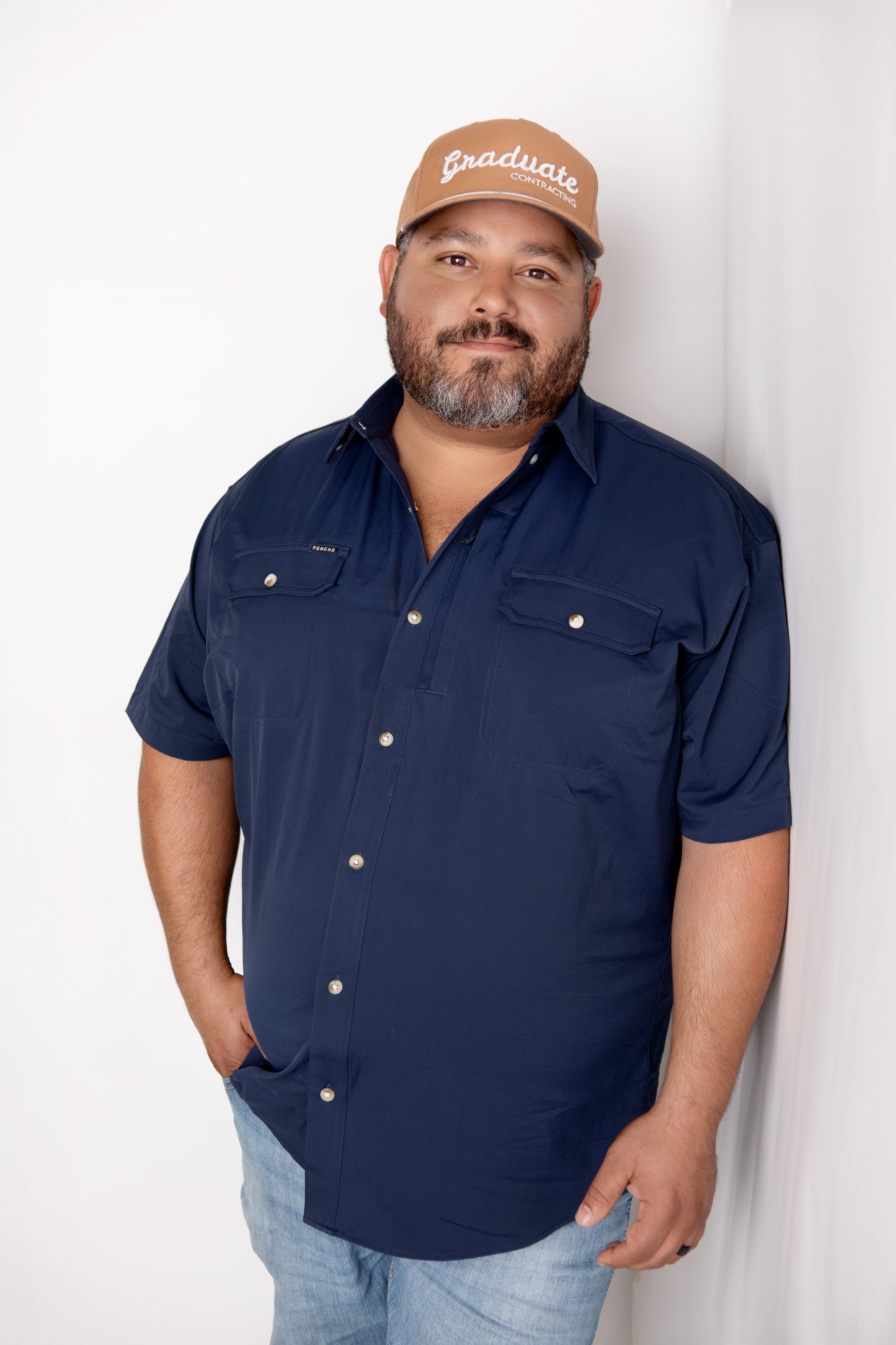 A man wearing a hat and a blue shirt is standing next to a wall.