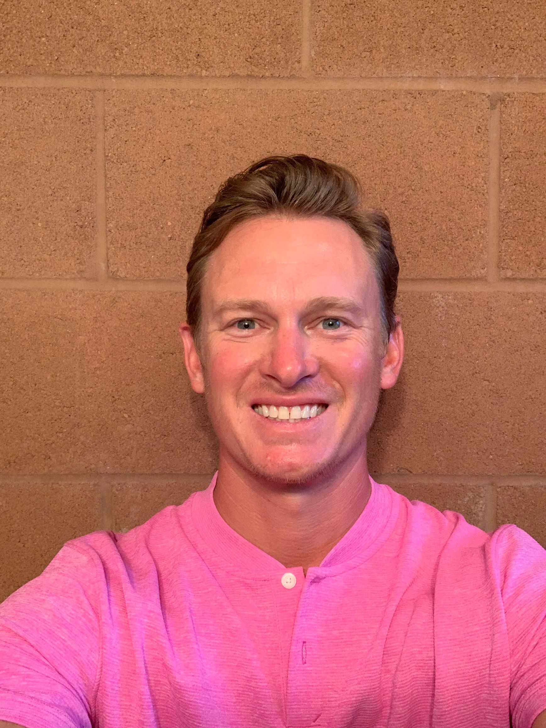 A man in a pink shirt is smiling in front of a brick wall.