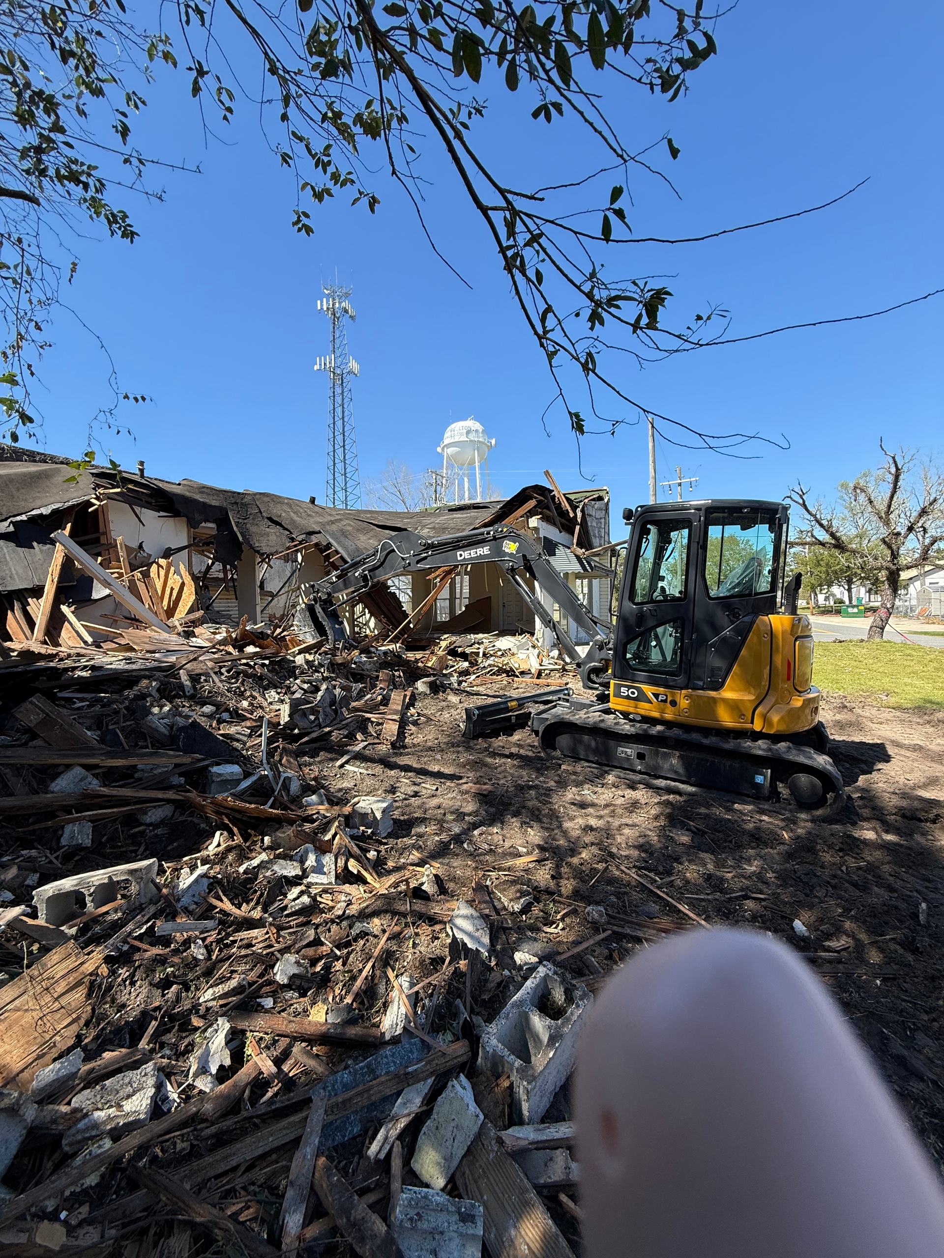 A yellow excavator demolishes a building, rubble pile in foreground, other buildings, tower, cloudy sky.